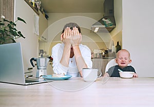 Young mother with coffee fighting tiredness while breakfast with baby in kitchen. Freelancer mom and child after sleepless night.