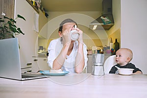 Young mother with coffee fighting tiredness while breakfast with baby in kitchen. Freelancer mom and child after sleepless night.