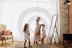 Young mother combing her little daughter`s hair standing in front of the mirror and her second daughter comes to them in