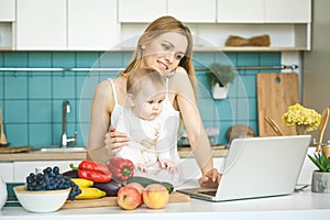 Young mother is cooking and playing with her baby daughter in a modern kitchen setting. Healthy food concept. Looking at laptop,