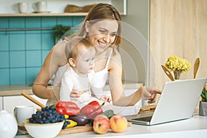 Young mother is cooking and playing with her baby daughter in a modern kitchen setting. Healthy food concept. Looking at laptop,