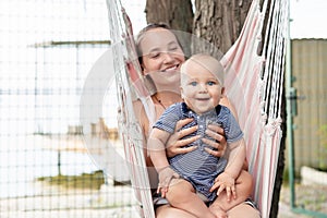 Young mother with cute baby boy sitting and relaxing on hammock near river or lake. Mom and kid having fun at summer outdoors. Hap