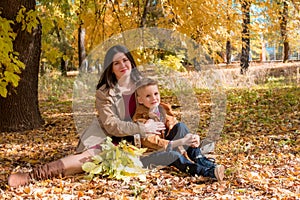 A young mother with dark hair in a beige autumn raincoat with her son in a sunny autumn park among yellow leaves. Autumn walk