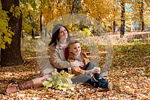 A young mother with dark hair in a beige autumn raincoat with her son in a sunny autumn park among yellow leaves. Autumn walk