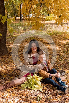 A young mother with dark hair in a beige autumn raincoat with her son in a sunny autumn park among yellow leaves. Autumn walk