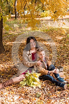 A young mother with dark hair in a beige autumn raincoat with her son in a sunny autumn park among yellow leaves. Autumn walk