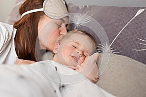 Young mother dark haired woman kissing sweet little baby, cute kid laying near mom and sleeping while parent kissing her, home