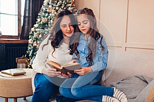Young mother and daughter reading book by Christmas tree in living room. Family spending time together at home