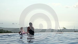 Young mother with daughter relaxing in infinity swimming pool looking at view