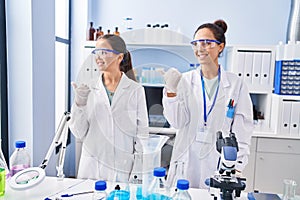 Young mother and daughter at scientist laboratory smiling with happy face looking and pointing to the side with thumb up