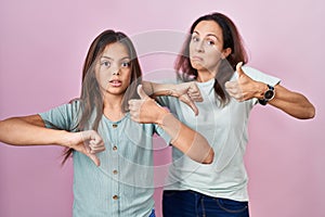 Young mother and daughter standing over pink background doing thumbs up and down, disagreement and agreement expression