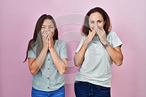 Young mother and daughter standing over pink background laughing and embarrassed giggle covering mouth with hands, gossip and