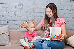 Young mother and daughter of two years old blonde use laptop computer laptop white with bright print sitting on couch indoors