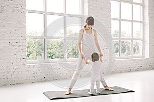 Young mother does physical yoga exercises together with her baby, mother and little child holding hands, standing on the