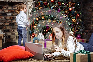 A young mother is doing online shopping with laptop and credit card while her daughter decorates the Christmas tree.