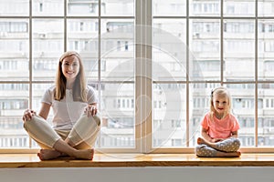 Young mother doing yoga with 3-years daughter in front of window. Happy mom smiling while practicing yoga together with