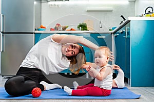 A young mother is doing yoga on a mat in the kitchen, and her baby is sitting next to her. The concept of home sports training