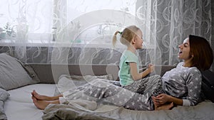 Young mother, dressed in pajamas and her daughter having fun on the bed in the bedroom, enjoying each other`s company