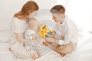 Young mother and father sit with little baby on bed, man give bouquet of yellow tulips on white background. Cute