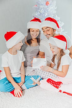 Young mother, father and two little children opening a magical Christmas gift by a Christmas tree in cozy living room in