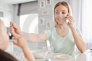 Young mother feeding child with spoon and talking on the phone