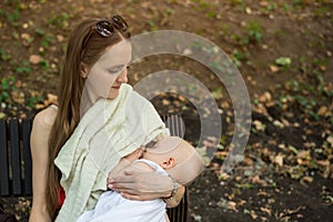 Young mother feeds the baby on a bench in the Park. Mom breastfeeding baby in public place