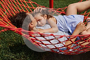 Young mother gently hugs her little daughter lying together on a hammock, enjoying the time in a place on a nature on a warm sunny