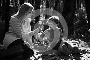 Young mother giving her little son a sweet season berrie at autumn picnic. Mum and kid sitting at picnic blanket and