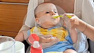 Young mother giving soup to hear little baby boy with plastic spoon in kitchen