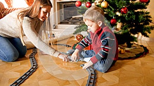 Young mother helping little son building tailways for toy train under Christmas tree at living room. Child receiving