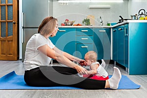 A young mother with her baby is doing yoga on a mat in the kitchen. The concept of home sports training with children