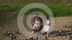 Young mother with her baby girl daughters feeding swan and little ducklings birds bread at a river wearing dotted dress