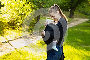 A young mother with her baby in a sling scarf is standing in a sunlight. Mom looks at the child. Wrapping sling for carrying a