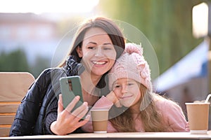 Young mother and her child daughter taking picture with phone selfie camera sitting at street cafe with hot drinks on