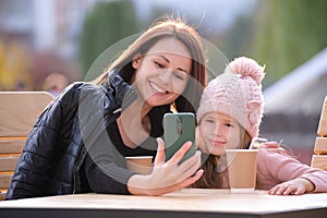 Young mother and her child daughter taking picture with phone selfie camera sitting at street cafe with hot drinks on