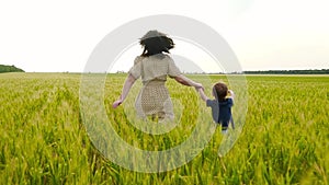 A young mother and her child are running across a field of green and yellow wheat, holding hands, in slow motion