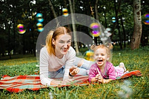 Young mother with her cute little girl is blowing a soap bubbles in summer park.