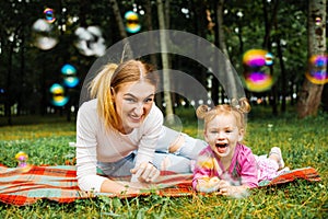 Young mother with her cute little girl is blowing a soap bubbles in summer park.