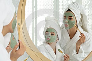 Young mother and her daughter with facial masks having fun near mirror in bathroom