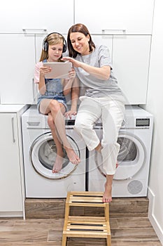 Young mother and her daughter are playing with tablet in laundry room sitting on washing machine