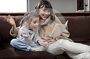 Young mother with her daughter are reading a book at home while sitting on the couch
