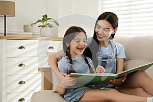 Young mother and her daughter reading book on sofa at home, space for text