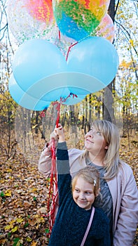 A young mother and her a little adorable daughter with the colorful balloons are walking in autumn forest.