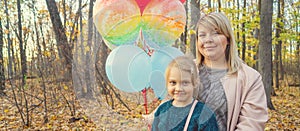 A young mother and her a little adorable daughter with the colorful balloons are walking in autumn forest.