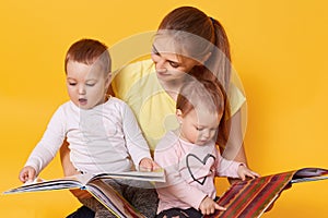 Young mother and her little babies daughters reading books, look on colourful pages, momny keeps children in her knees while