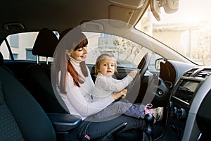 Young mother with her little baby girl sitting in the car in the front seat near car steering wheel. A woman and baby