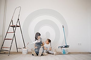 Young mother and her little blonde cute girl with wall paint rollers and brushes sitting on floor