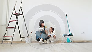 Young mother and her little blonde cute girl with wall paint rollers and brushes sitting on floor