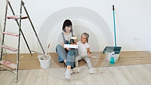 Young mother and her little blonde cute girl with wall paint rollers and brushes sitting on floor