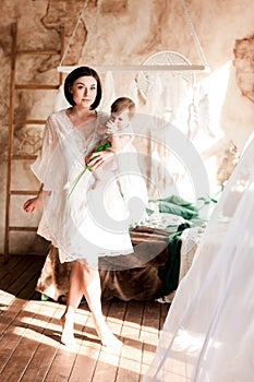 Young mother with her little daughter in her arms opposite the window with a white curtain